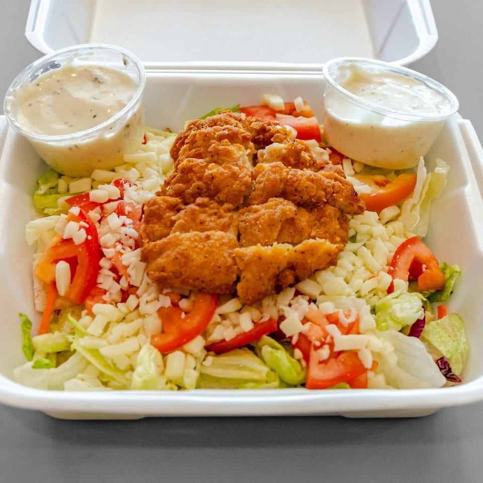 Crispy chicken salad with shredded cheese, tomatoes, and two dressing cups in a white takeout container.