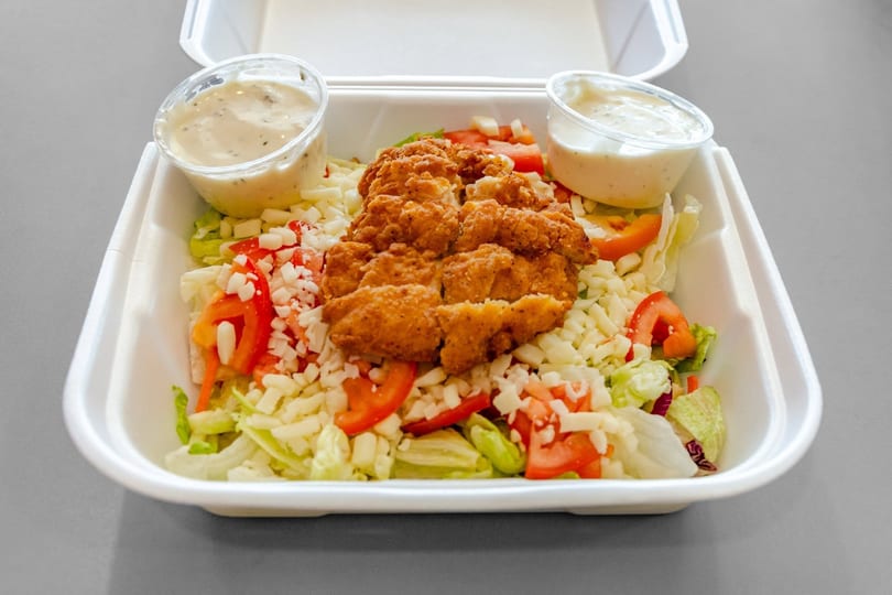 Crispy chicken salad with shredded cheese, tomatoes, and two dressing cups in a white takeout container.
