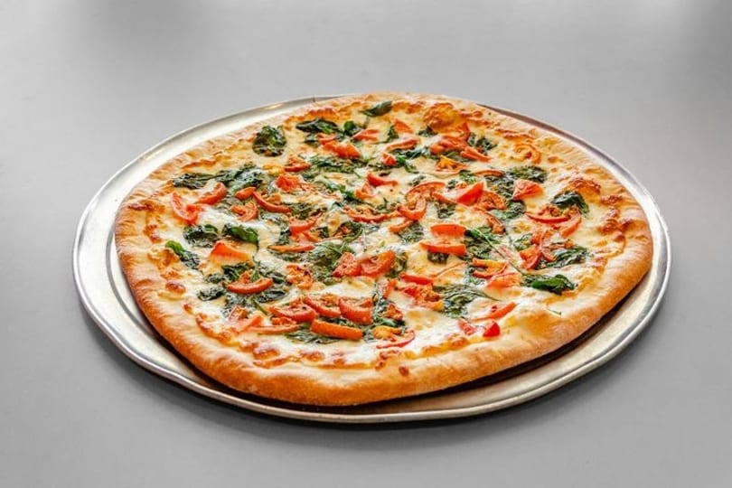 A whole pizza with melted cheese, spinach, and diced tomatoes on a metal tray.